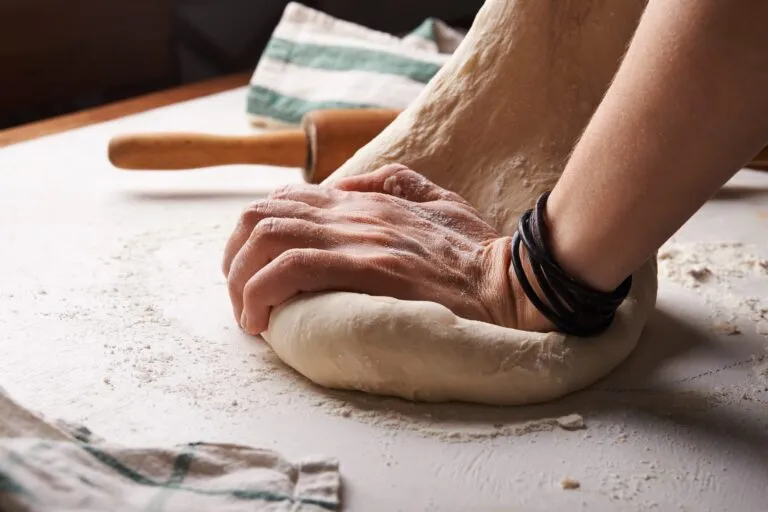 Hände kneten Pizzateig auf einer bemehlten Arbeitsfläche, daneben ein Nudelholz